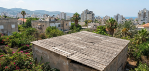מבנה שירות ישן בעל גג אסבסט צמנט גלי ובלוי הנמצא בחצר בשכונת מגורים בנהריה, מוקף בצמחייה ירוקה, כשברקע נראים בנייני מגורים עירוניים. התמונה מדגימה מבנה המהווה מפגע פוטנציאלי .